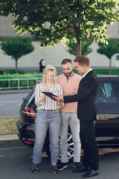 Un giovane venditore mostra ai clienti un'auto nuova. Coppia, uomo e donna felici acquistano una nuova auto. I giovani firmano documenti per acquistare un'auto.