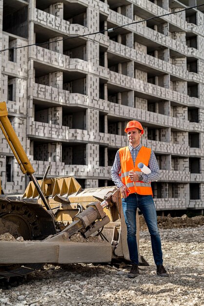 Un grande lavoratore in uniforme e casco sta accanto a un escavatore in un cantiere edile Un escavatore è in costruzione Concetto di costruzione