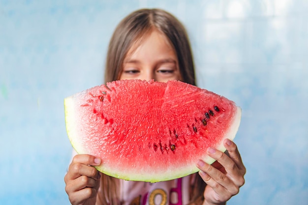 Un grande pezzo di anguria rossa matura nelle mani di una bambina su sfondo blu bambino di 8 anni che mangia anguria a casa