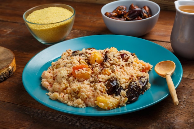 Un grande piatto di cuscus su un tavolo di legno accanto ai cereali in una ciotola di noci e una brocca di brodo di frutta