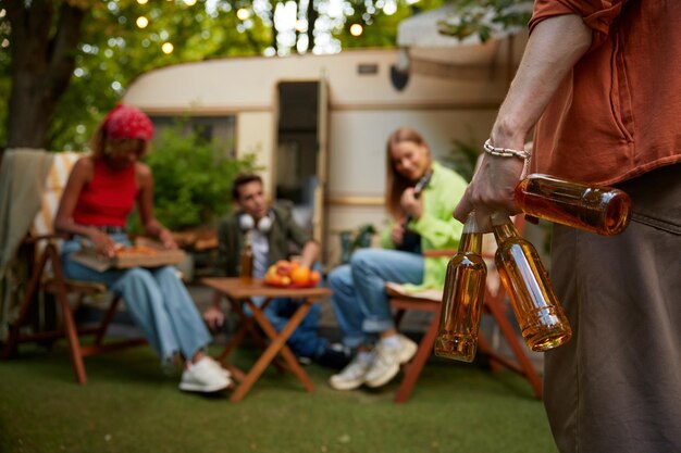 Un gruppo di amici felici che si riposano in campeggio vicino al loro camper nella foresta. Concentrati sulla mano maschile che tiene diverse bottiglie di birra appena raffreddata