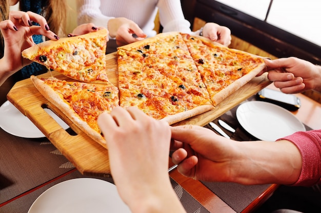 Un gruppo di amici prende una pizza calda e fresca seduti in un caffè. Mani con fette di pizza close-up.