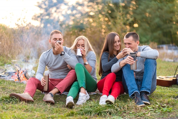 Un gruppo di amici si sta godendo una bevanda calda da un thermos, in una fresca serata accanto a un incendio nella foresta. Divertimento in campeggio con gli amici