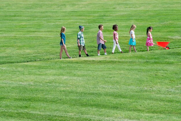 Un gruppo di bambini che cammina su un sentiero in pendenza in ordine di altezza seguendo il più piccolo davanti con una carriola rossa