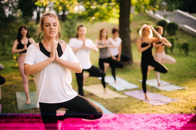 Un gruppo di donne yogini sono in piedi su stuoie di yoga e si equilibrano su una gamba in un albero di posa nel parco in una mattina di sole estivo con l'allenatore. Un gruppo di persone sta praticando yoga in piedi nell'esercizio Vrksasana