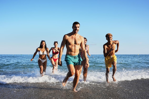 Un gruppo di giovani amici felici in costume da bagno stanno correndo fuori dall'acqua verso la spiaggia