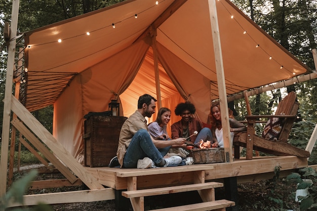 Un gruppo di giovani sorridenti che arrostiscono marshmallow su spiedini sul pozzo del fuoco al campeggio, godendosi una vacanza glamping all'aperto con gli amici insieme riaprono dopo il blocco della pandemia.