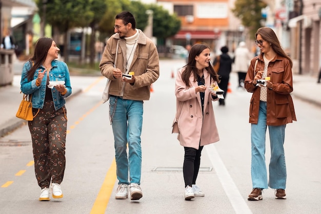 Un gruppo di giovani vestiti con abiti moderni parla mentre passeggia per la città e mangia dolci Messa a fuoco selettiva