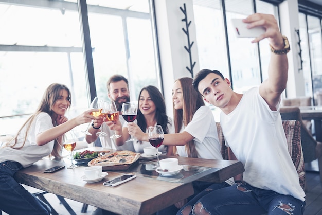 Un gruppo di persone fa una foto selfie in un caffè. I migliori amici si sono riuniti a tavola per mangiare la pizza e cantare vari drink