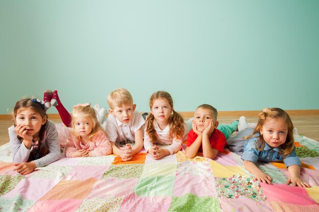 Un gruppo di sei graziosi bambini, ragazzi e ragazze di diverse età, sdraiati sul pavimento sulla coperta patchwork colorata a casa in soggiorno guardando la fotocamera con interesse