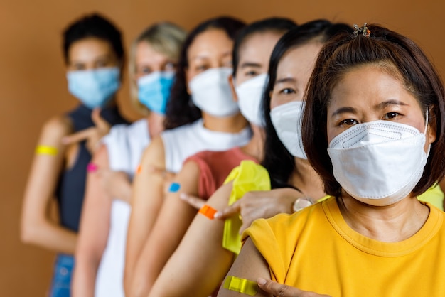 Un gruppo eterogeneo di pazienti di sesso femminile etnico multinazionale indossa una maschera facciale in fila per ordine di altezza guarda la telecamera che punta insieme cerotti colorati sulla spalla dopo la vaccinazione.