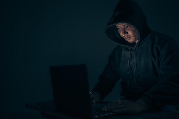 Un hacker con un cappuccio che gli copre la testa Seduto in una stanza buia usando un laptop