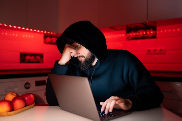 Un hacker con una felpa con cappuccio nera con un cappuccio in testa è seduto davanti a un laptop e si tiene tristemente sul viso