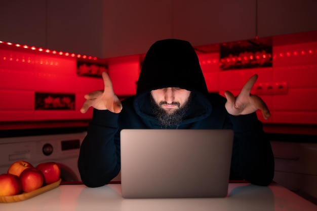 Un hacker con una felpa con cappuccio nera e un cappuccio in testa è seduto davanti a un laptop che mostra il pollice in alto e si rallegra per un hack riuscito