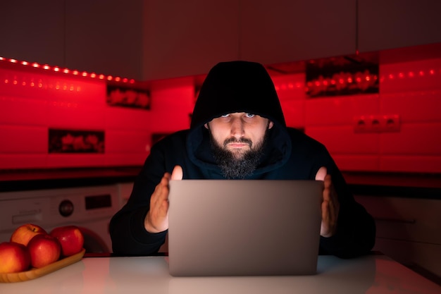 Un hacker con una felpa con cappuccio nera e un cappuccio in testa è seduto davanti a un laptop con in mano il coperchio e guarda con sospetto