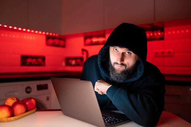 Un hacker con una felpa con cappuccio nera e un cappuccio in testa è seduto davanti a un laptop proteso in avanti e guarda attentamente
