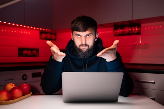 Un hacker con una felpa nera con cappuccio è seduto davanti a un laptop che mostra i palmi delle mani in avanti