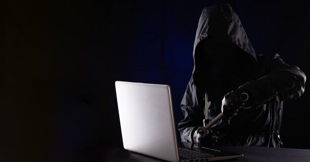Un hacker criminale in un cappuccio in abiti neri e passamontagna distrugge un laptop con strumenti, una chiave inglese su sfondo nero