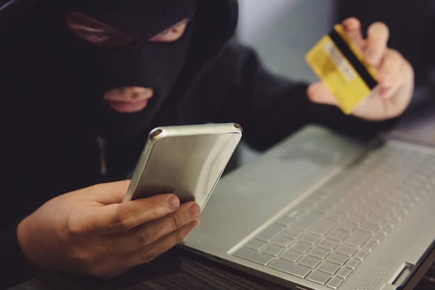 Un hacker maschio con una maschera da rapinatore utilizza telefono, carta di credito e laptop in uno schema fraudolento. Il ladro informatico ha rubato i dati personali e le informazioni sulla carta di credito. L'hacker utilizza il malware per rubare i soldi degli utenti.