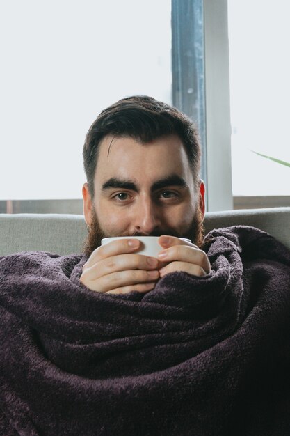 Un hipster giovane con la barba che tiene una tazza di caffè mentre è coperto su una coperta nel divano