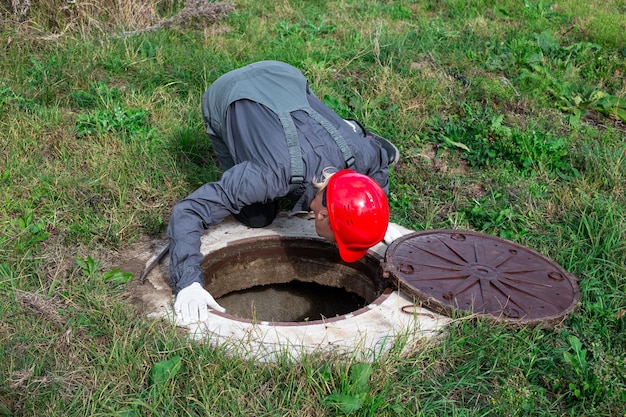 Un idraulico maschio ha aperto il portello di un pozzo d'acqua e guarda all'interno Ispezione dei tubi dell'acqua e dei contatori