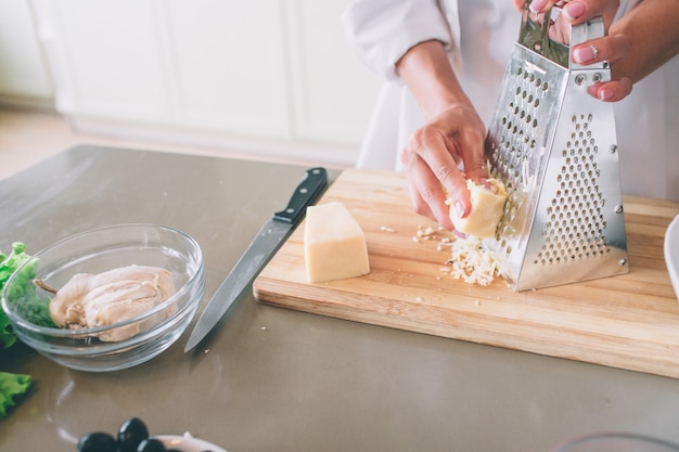 Un'immagine delle mani della donna che grattano pezzo di formaggio sulla grattugia. È su tavola di legno. Il coltello è sdraiato sul tavolo.