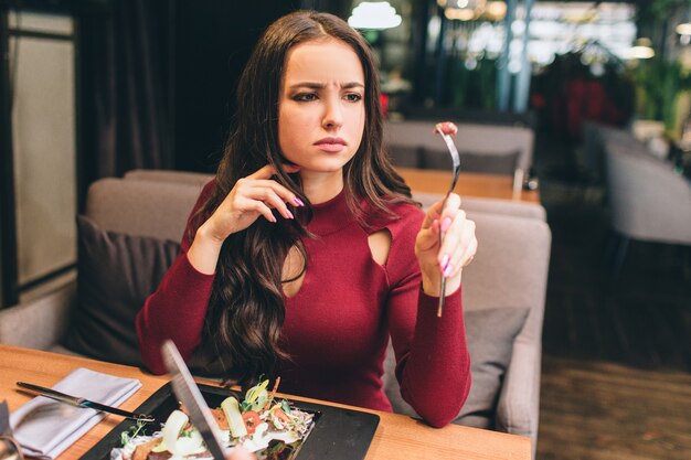 Un'immagine di bella donna che si siede alla tavola in ristorante e che mangia la sua insalata. Fece un incontro sulla forchetta e lo guardò. È preoccupata per qualcosa.