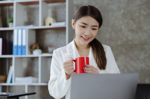 Un'impiegata asiatica si siede con una tazza di caffè rossa nell'ufficio del dipartimento marketing della startup, è un collegamento con i clienti di marketing e un'operaia di consulenza Concetto di marketing