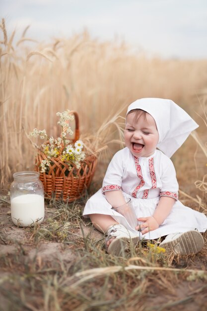 Un'infanzia sana in armonia con la natura. piccolo bambino felice in abiti popolari si siede su un campo di grano con un barattolo di latte fresco