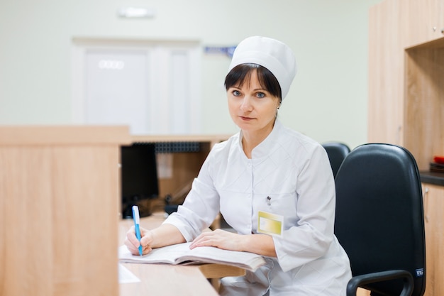 Un'infermiera in un camice medico bianco siede a una scrivania di legno alla reception e fa le voci in un registro di lavoro. Donna medico che lavora