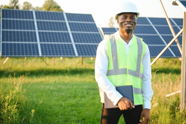 Un ingegnere afroamericano con una camicia bianca e un elmetto sta lavorando su un campo di pannelli solari Concetto di energia solare rinnovabile