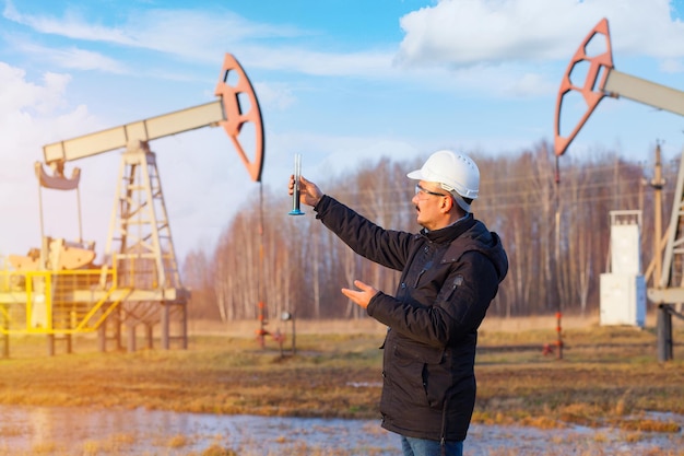 Un ingegnere chimico controlla la qualità dell'olio estratto. Un uomo con un casco protettivo bianco tiene una provetta con petrolio greggio sullo sfondo di una sedia a dondolo a olio