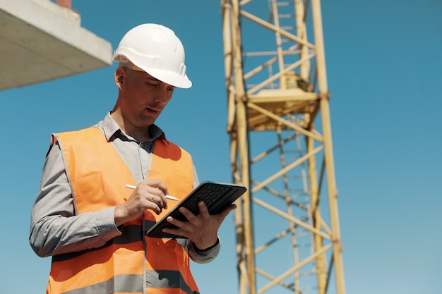 Un ingegnere con un giubbotto arancione e un casco bianco per il controllo della costruzione conduce un'ispezione con un tablet in mano contro il lato di un cantiere e una gru a torre