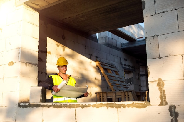 Un ingegnere edile e designer con un elmetto protettivo giallo studia il disegno di un edificio in un cantiere edile. Una casa fatta di blocchi di cemento porosi, un lavoratore nel processo alla finestra e al muro