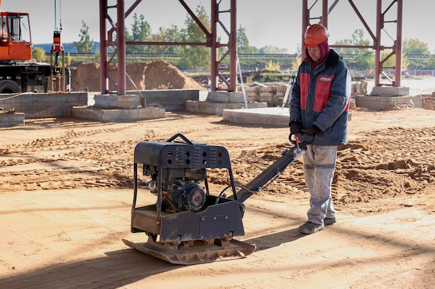 Un lavoratore compatta la terra con un costipatore vibrante in un cantiere in una luminosa giornata di sole. Preparazione della superficie per l'installazione di un pavimento in cemento.