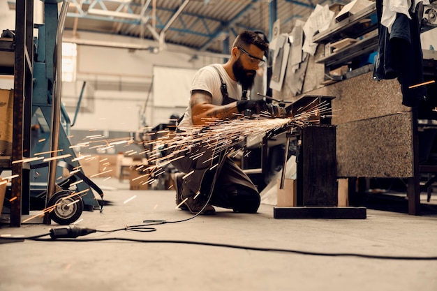 Un lavoratore dell'industria pesante inginocchiato in una struttura e che lavora parti metalliche con una smerigliatrice