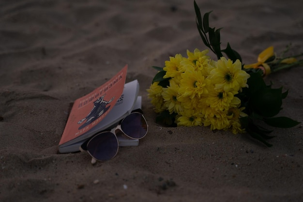 Un libro con occhiali da sole e un mazzo di fiori gialli si trova su una spiaggia sabbiosa