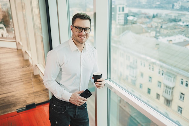 Un manager di sesso maschile con una camicia bianca e occhiali si trova nell'ufficio vicino alla finestra e beve caffè mentre guarda un video al telefono Conversazione telefonica d'affari di un uomo d'affare