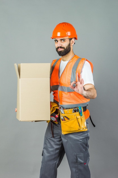Un manbuilder in un casco arancione con una scatola di cartone nelle sue mani su uno sfondo grigio