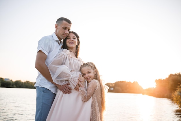 Un marito con una moglie e una figlia incinta sta riposando sulla riva del fiume in piedi sul molo in estate tempo soleggiato al tramonto coppia innamorata uomo e donna che si abbracciano tenendosi per mano Una famiglia felice