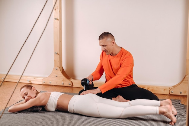 Un massaggiatore maschio massaggia il corpo di una ragazza con un massaggiatore a percussione per prepararsi a un simulatore di stretching