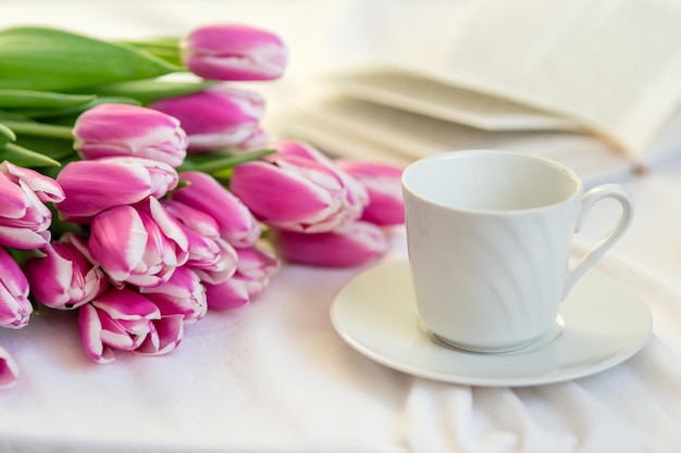 Un mazzo di bei tulipani rosa giace su un letto bianco accanto a una tazza e un piattino bianchi e un libro aperto