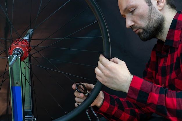 Un meccanico di biciclette con una camicia a quadri rossa sta assemblando una ruota utilizzando un supporto per centraggio professionale su sfondo nero Riparazione di biciclette Il viso e le mani del meccanico sono in primo piano durante il lavoro