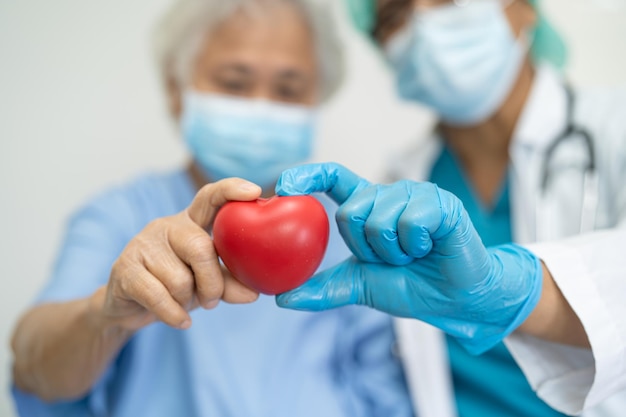 Un medico che indossa un ppe con una maschera dà un cuore rosso per proteggere il coronavirus a una paziente asiatica anziana o anziana.