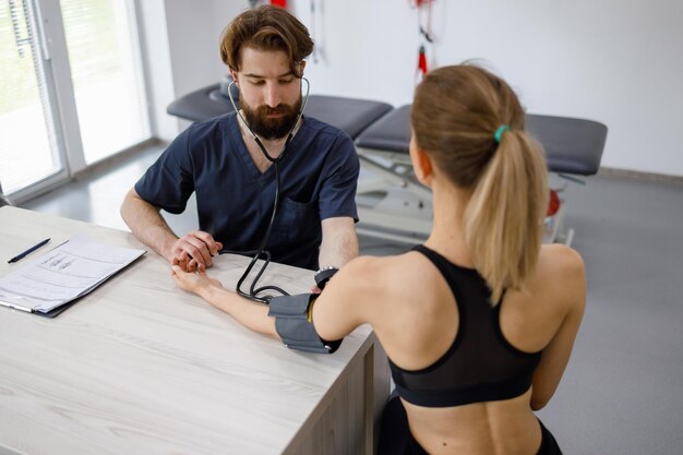 Un medico fisioterapista misura la pressione di una giovane donna in forma seduta in una palestra di riabilitazione