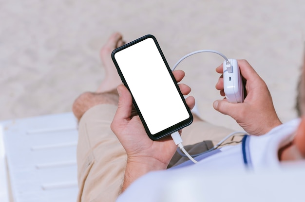 Un mock-up di uno smartphone nelle mani di un ragazzo con un caricabatterie di Power Bank. Sullo sfondo della spiaggia di sabbia.