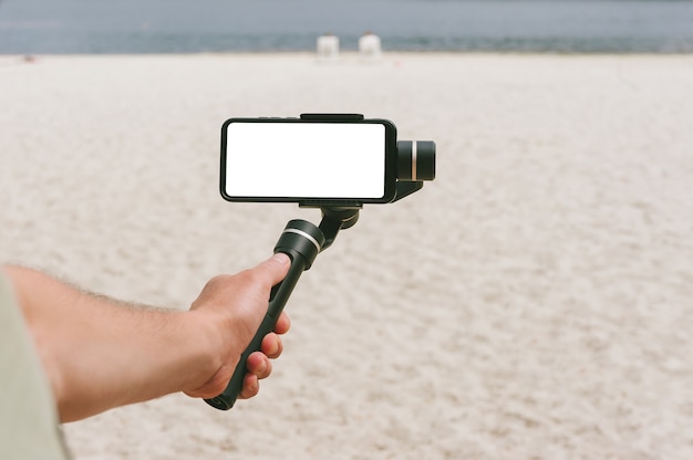 Un mock-up di uno smartphone su una Steadicam nella mano di un uomo. Sullo sfondo di una spiaggia di sabbia.