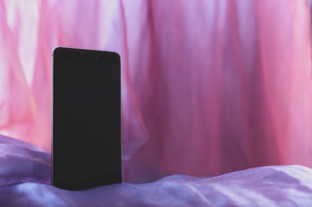 Un mockup con un cellulare nero in piedi lateralmente su un arioso panno rosa. Schermo nero con copia spazio. Lo sfondo è sfocato. Orizzontale.