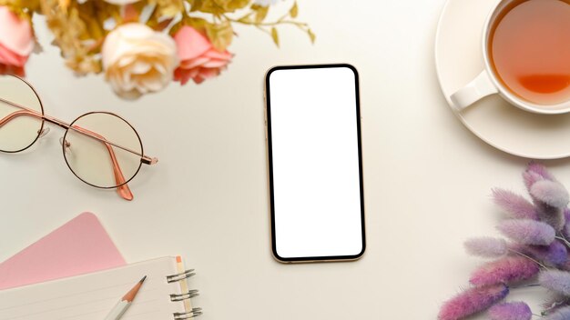 Un mockup vuoto dello schermo dello smartphone con decorazioni femminili su un'area di lavoro bianca con vista dall'alto piatta