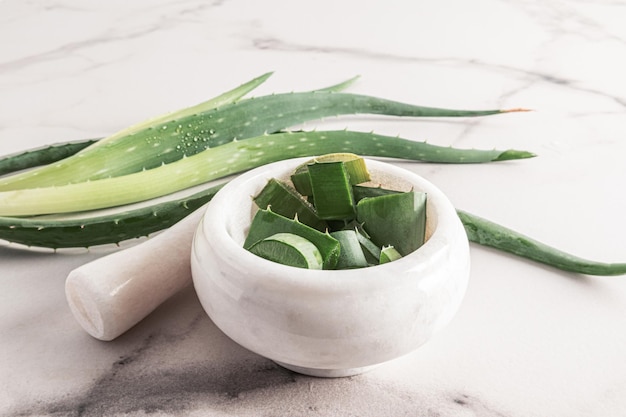 Un mortaio di marmo bianco con un pestello con fette di aloe vera affettate su uno sfondo di marmo accanto alle foglie fresche della pianta sullo sfondo di marmo